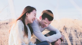 A photo of an Asian woman and Western man heartily laughing while sitting next to each other in a grass field