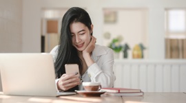 A photo of a foreign woman smiling while looking at her phone, with a laptop and cup of coffee in front of her